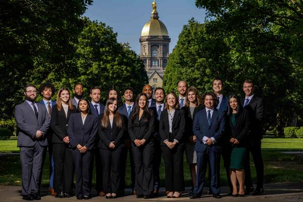 A group of nineteen students in business attire stand for a photo on campus, with the Main Building and its Golden Dome visible in the background.