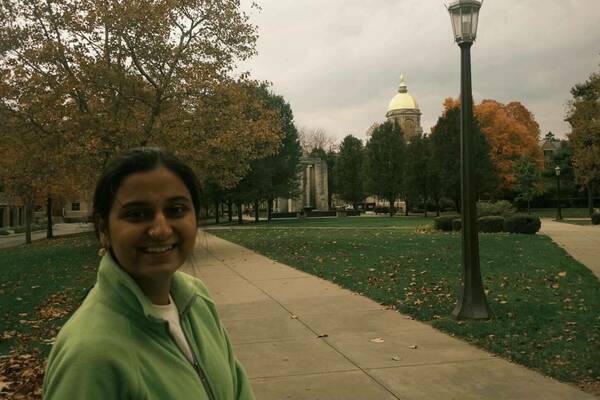 A smiling woman in a light green fleece stands on a Notre Dame campus path. The Golden Dome of the Main Building is visible in the autumn background, surrounded by trees with yellow and orange leaves, and grass scattered with brown leaves under a cloudy sky.