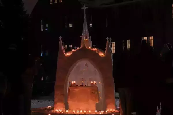 A detailed snow replica of the Grotto of Our Lady of Lourdes at Notre Dame, illuminated by many warm candles at night. Silhouettes of people observe the glowing snow chapel, with a dark campus building in the background.