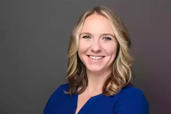 A woman with white skin and shoulder-length blonde hair poses with her arms crossed for a portrait. She is smiling and wearing a dark blue blouse.