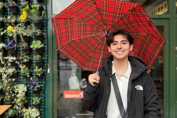 Smiling person holds a red plaid umbrella outside 'The Goodman's,' next to a green living wall and a wooden bench.