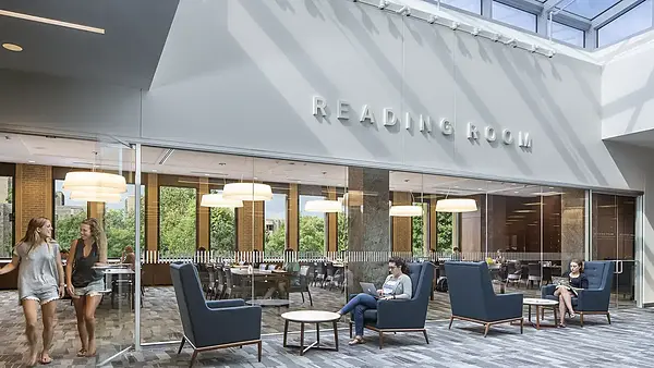 A brightly lit modern study area in Hesburgh Library with large lettering mounted on the wall above that reads "READING ROOM." Behind glass walls, students are seated at tables near large windows. In the foreground, outside of the reading room, a student works on a laptop in a blue high-backed armchair. On the left, two young women are walking through the reading room entrance.