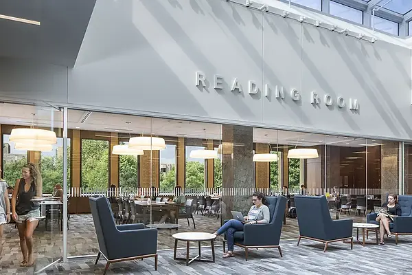 A brightly lit modern study area in Hesburgh Library with large lettering mounted on the wall above that reads "READING ROOM." Behind glass walls, students are seated at tables near large windows. In the foreground, outside of the reading room, a student works on a laptop in a blue high-backed armchair. On the left, two young women are walking through the reading room entrance.